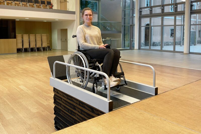 Hublift hebt einen Rollstuhl samt Nutzerin auf eine höhere Ebene